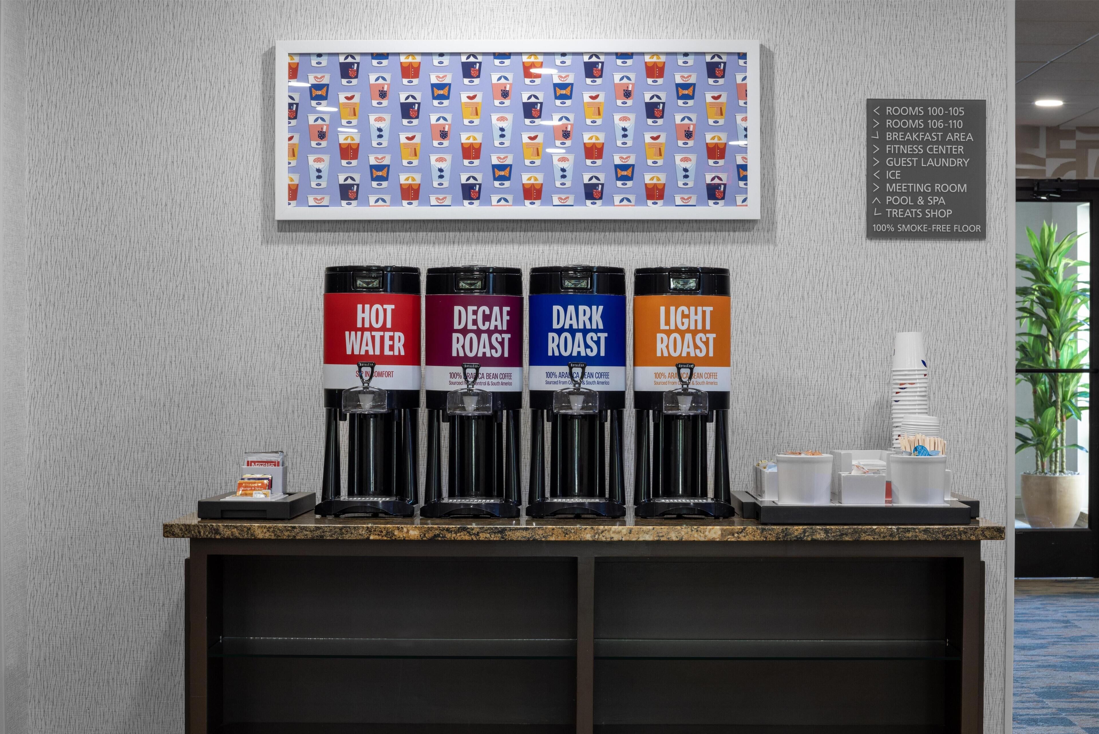 Ein Kaffee-Bereich mit vier Spendern, beschriftet mit 'Heißwasser', 'Entkoffeiniert', 'Dunkle Röstung' und 'Helle Röstung'. Darauf befindet sich eine Wandtafel, die Hoteleinrichtungen wie Zimmer, Frühstücksbereich, Fitnesscenter und weitere Angebote auflistet.