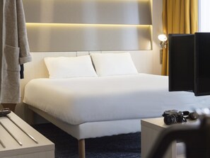 A hotel room with a large bed, a bedside table with a camera, a lamp, and a curtain. - ibis Styles Nivelles (Nivelles)