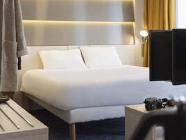 Una habitación de hotel con una cama grande, una mesita de noche con una cámara, una lámpara y una cortina.