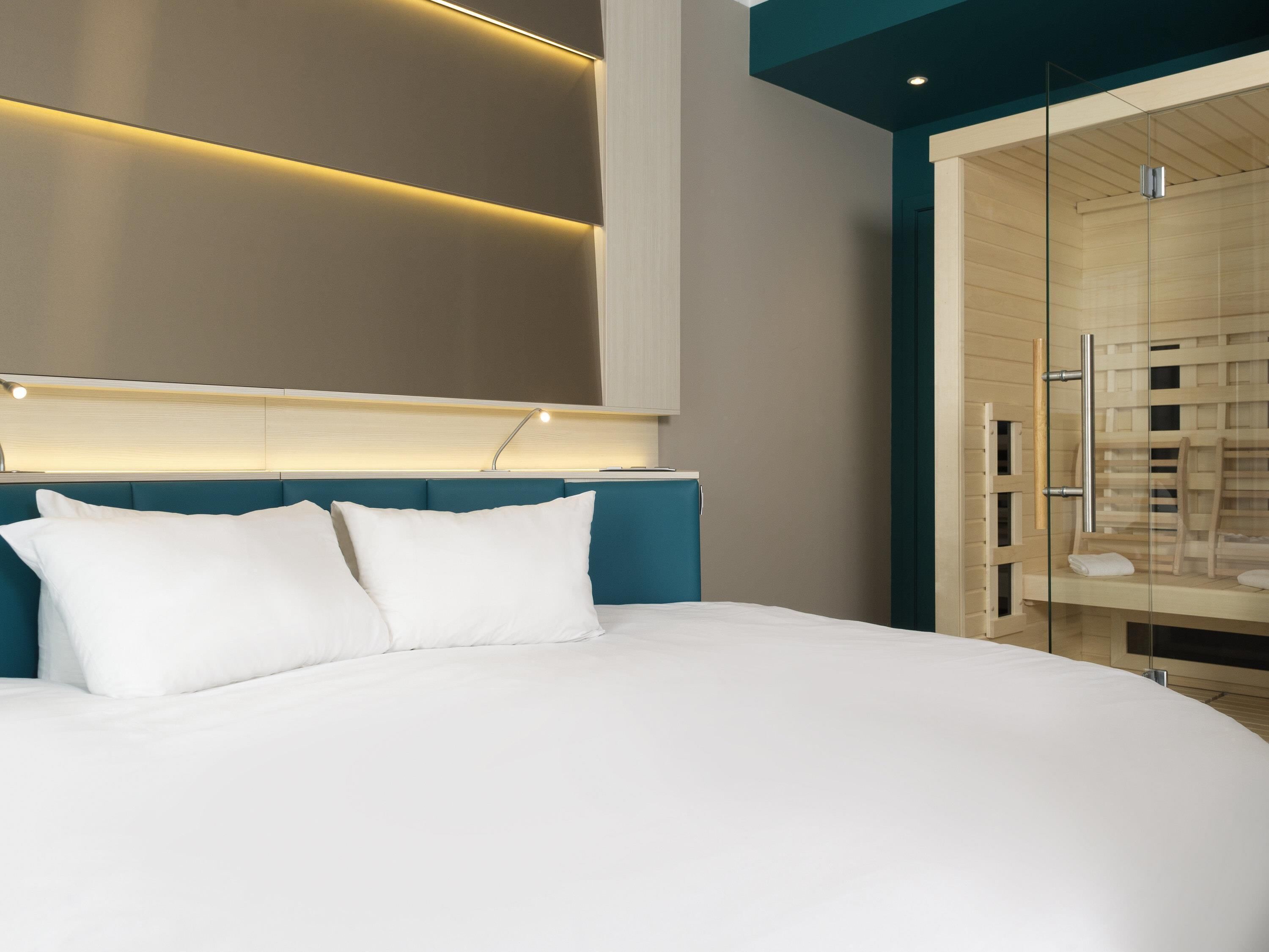 Habitación de hotel moderna con una cama grande, cabecera con iluminación integrada y zona de ducha con paredes de vidrio.