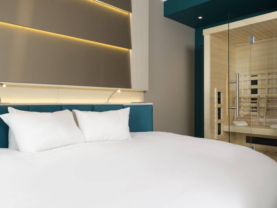 Une chambre d’hôtel moderne dotée d’un grand lit, d’une tête de lit avec éclairage intégré et d’une douche séparée par des parois en verre.
