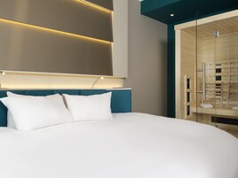 Habitación de hotel moderna con una cama grande, cabecera con iluminación integrada y zona de ducha con paredes de vidrio.