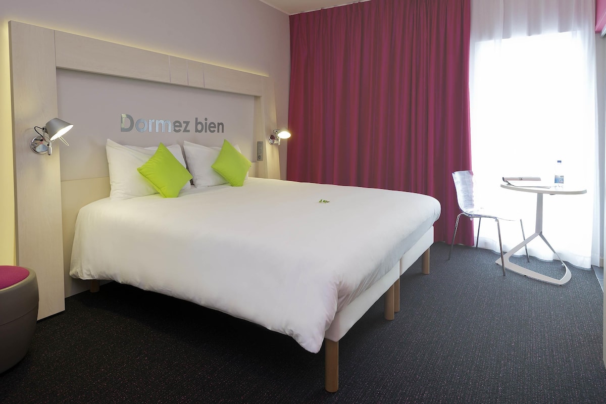 Habitación de hotel con cama, escritorio, silla y ventana con cortinas.