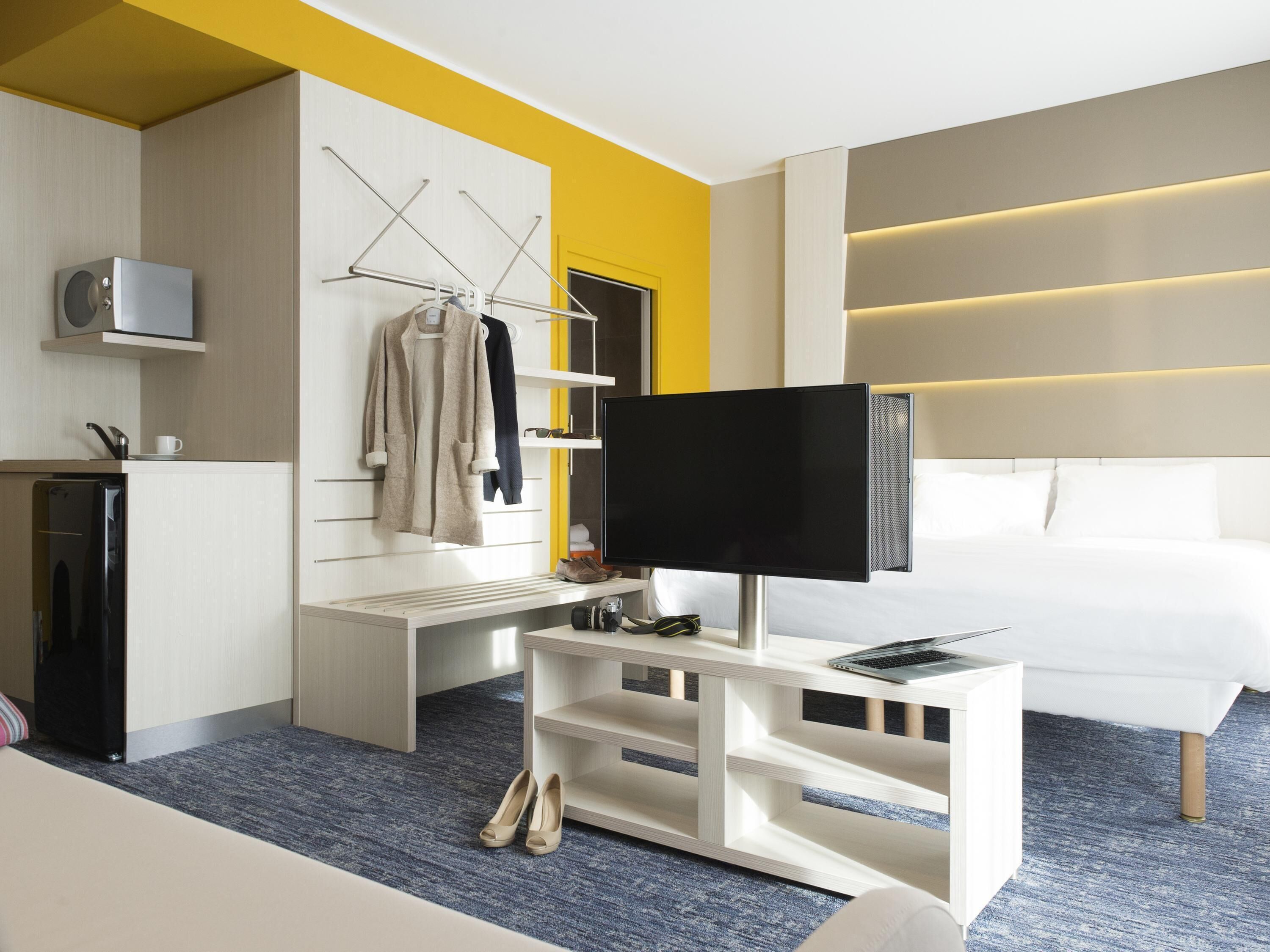 A modern hotel room with a flat-screen TV, a white sofa, a small kitchenette, and a seating area with a bench.