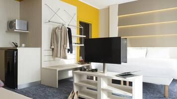 A modern hotel room with a flat-screen TV, a white sofa, a small kitchenette, and a seating area with a bench.