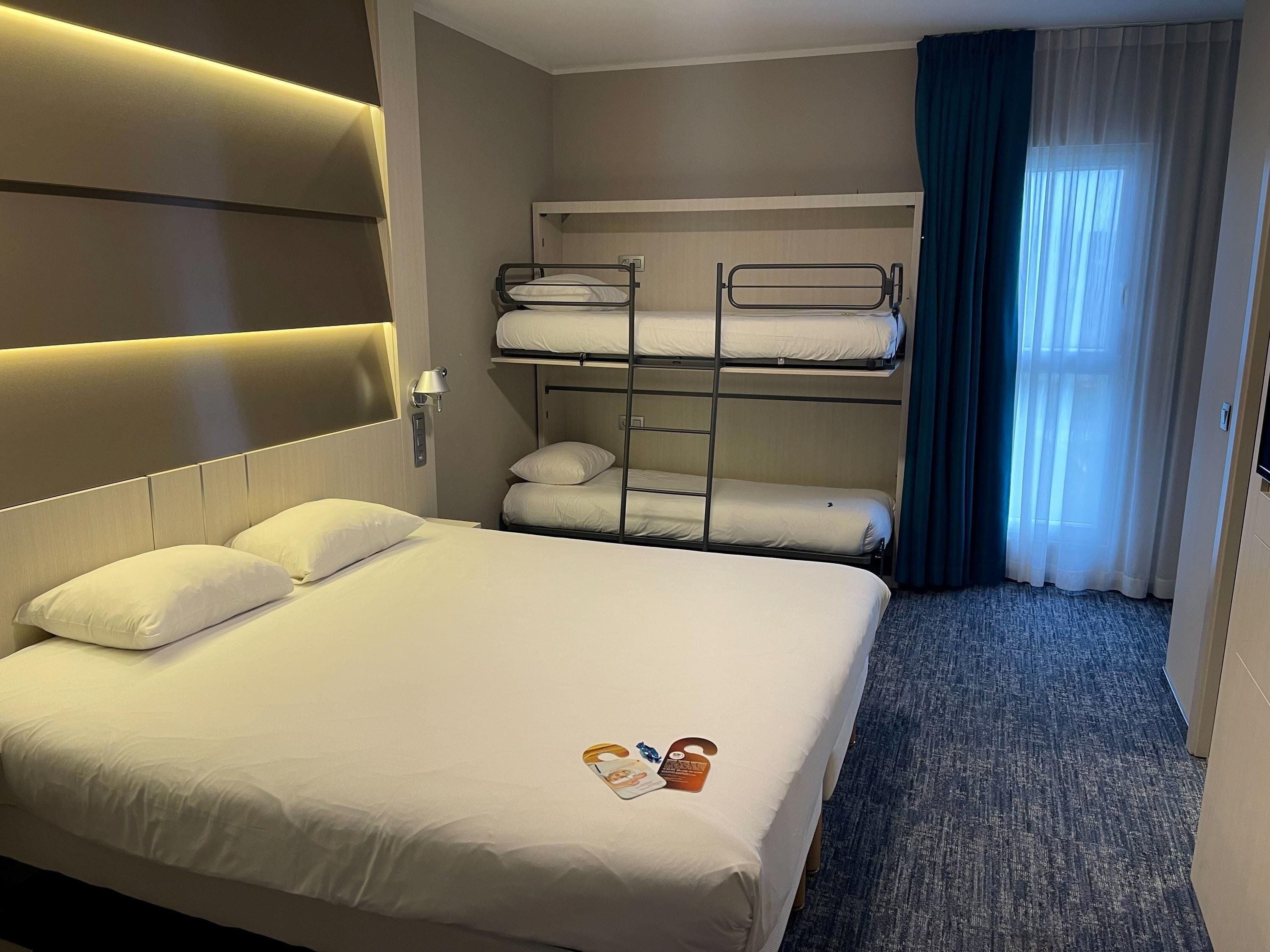Ein Hotelzimmer mit einem großen Bett, einem Stockbett, einer Nachttischlampe, einem Fenster mit Vorhängen und einem Teppichboden.