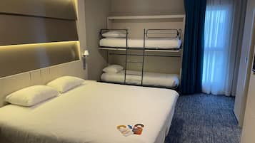 Ein Hotelzimmer mit einem großen Bett, einem Stockbett, einer Nachttischlampe, einem Fenster mit Vorhängen und einem Teppichboden.
