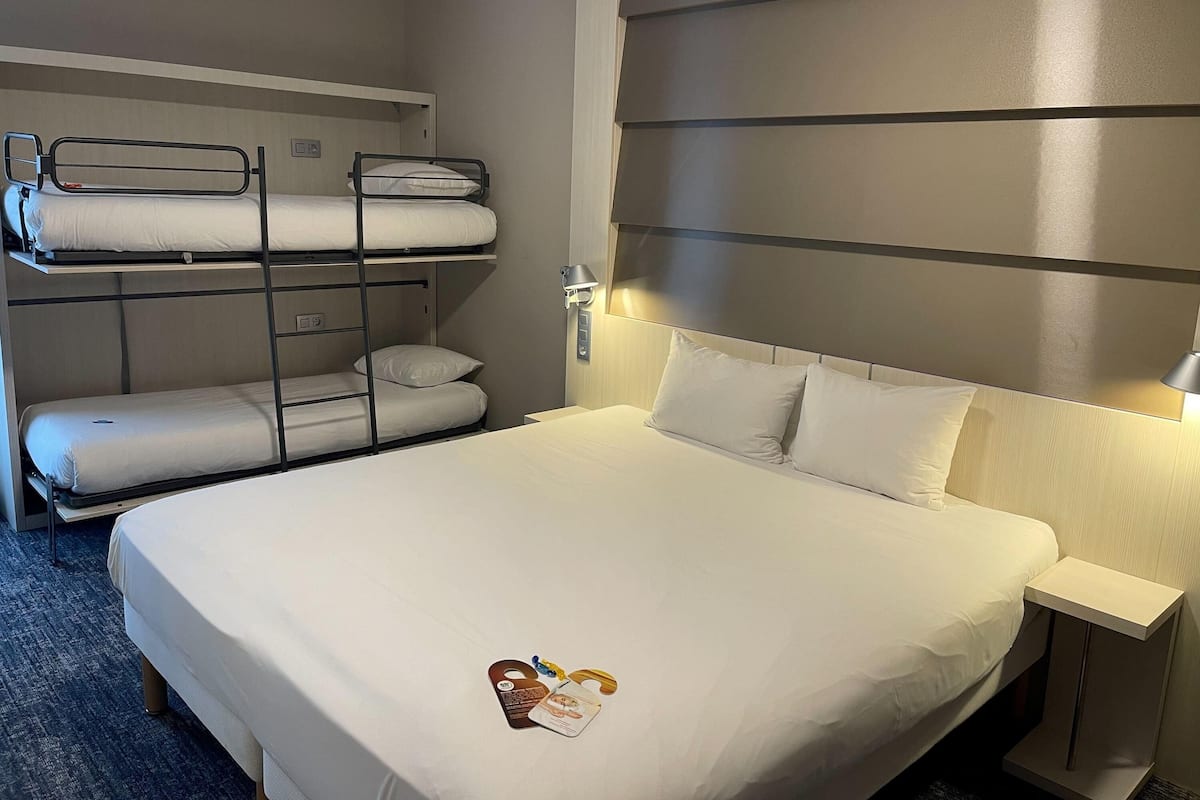 A hotel room with a bunk bed, a single bed, and a bedside table with a lamp.