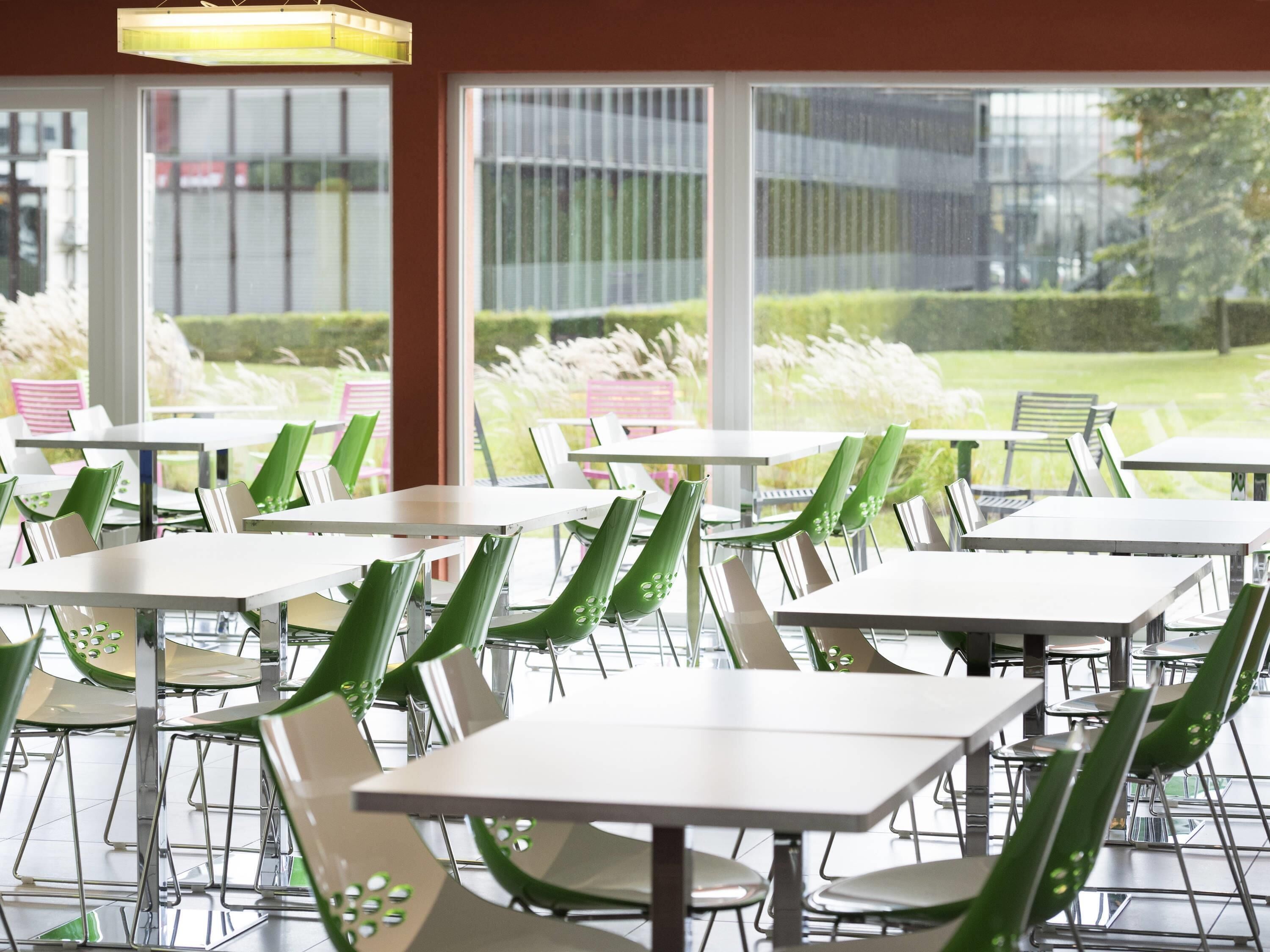 A restaurant with multiple tables and chairs, large windows, and a view of greenery outside.