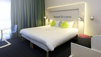 A hotel room with a bed, a chair, a small table, and a trash bin.