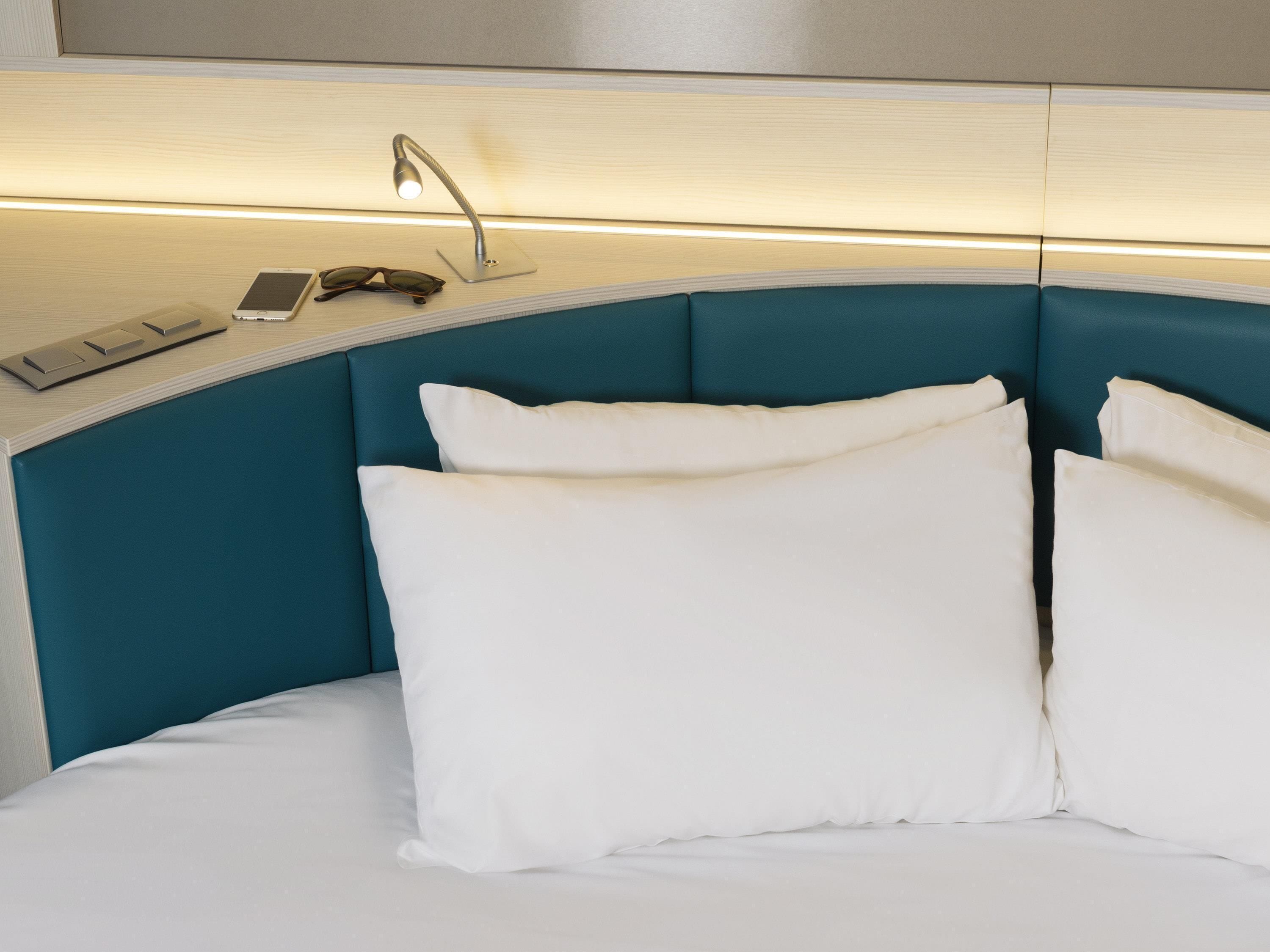 A neatly made bed with white bedding, a bedside lamp, a smartphone, and a pair of glasses on a bedside table.