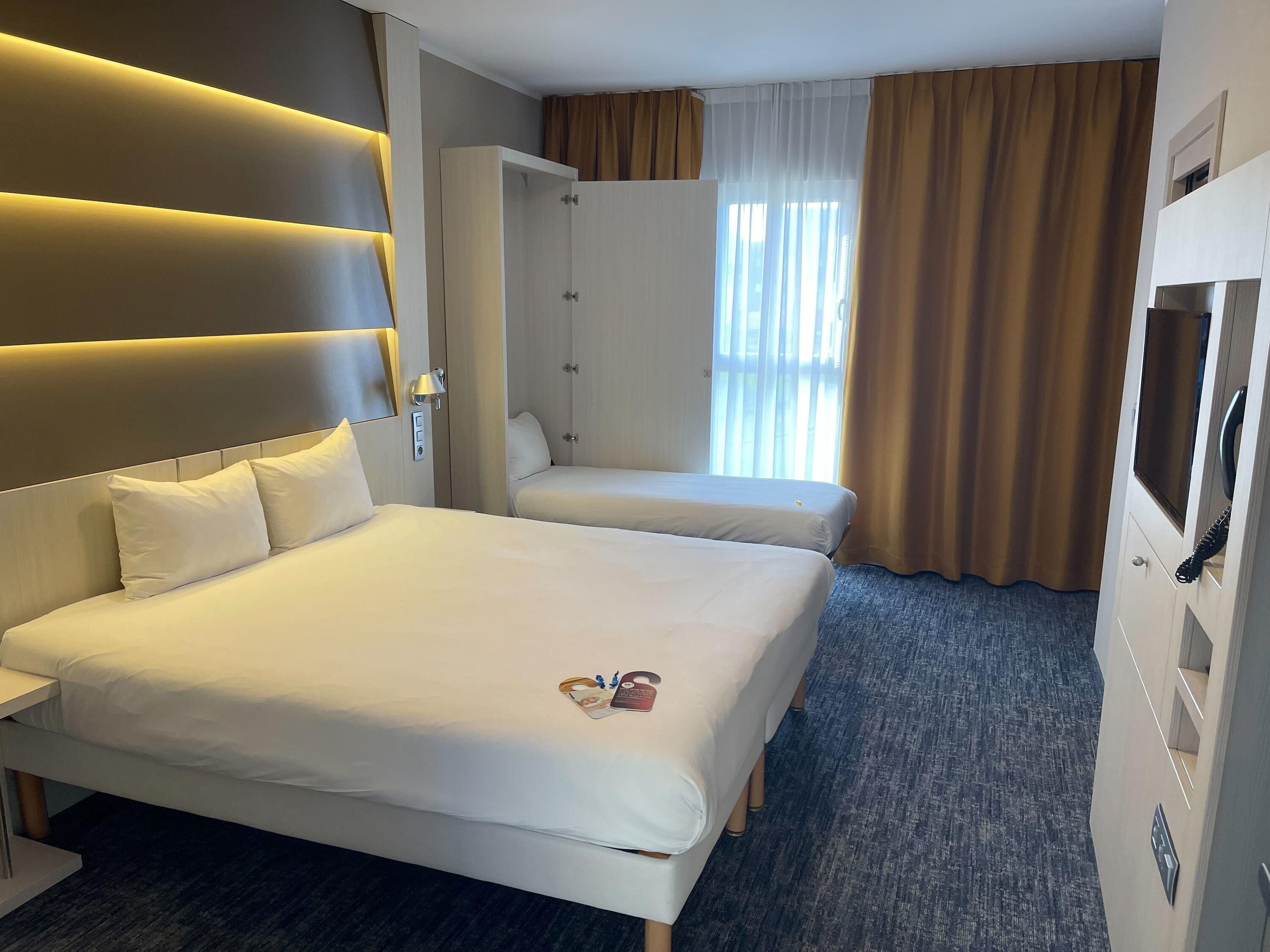 Una habitación de hotel con una cama grande, una cama más pequeña, un armario, un escritorio y un televisor.