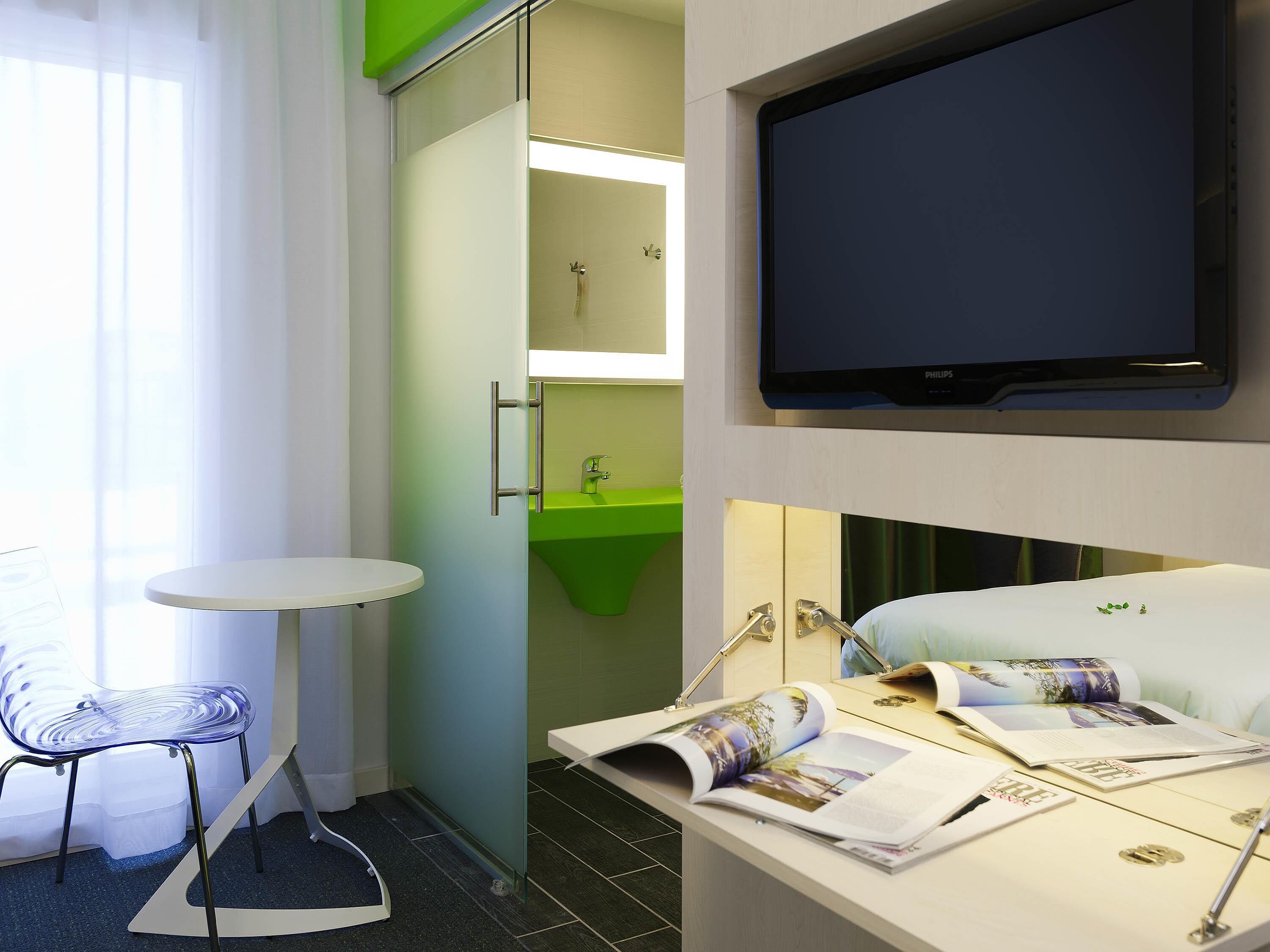Une chambre d’hôtel avec un lit, une télévision, une petite table, une chaise, un lavabo vert et un miroir.