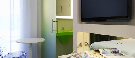 Une chambre d’hôtel avec un lit, une télévision, une petite table, une chaise, un lavabo vert et un miroir.