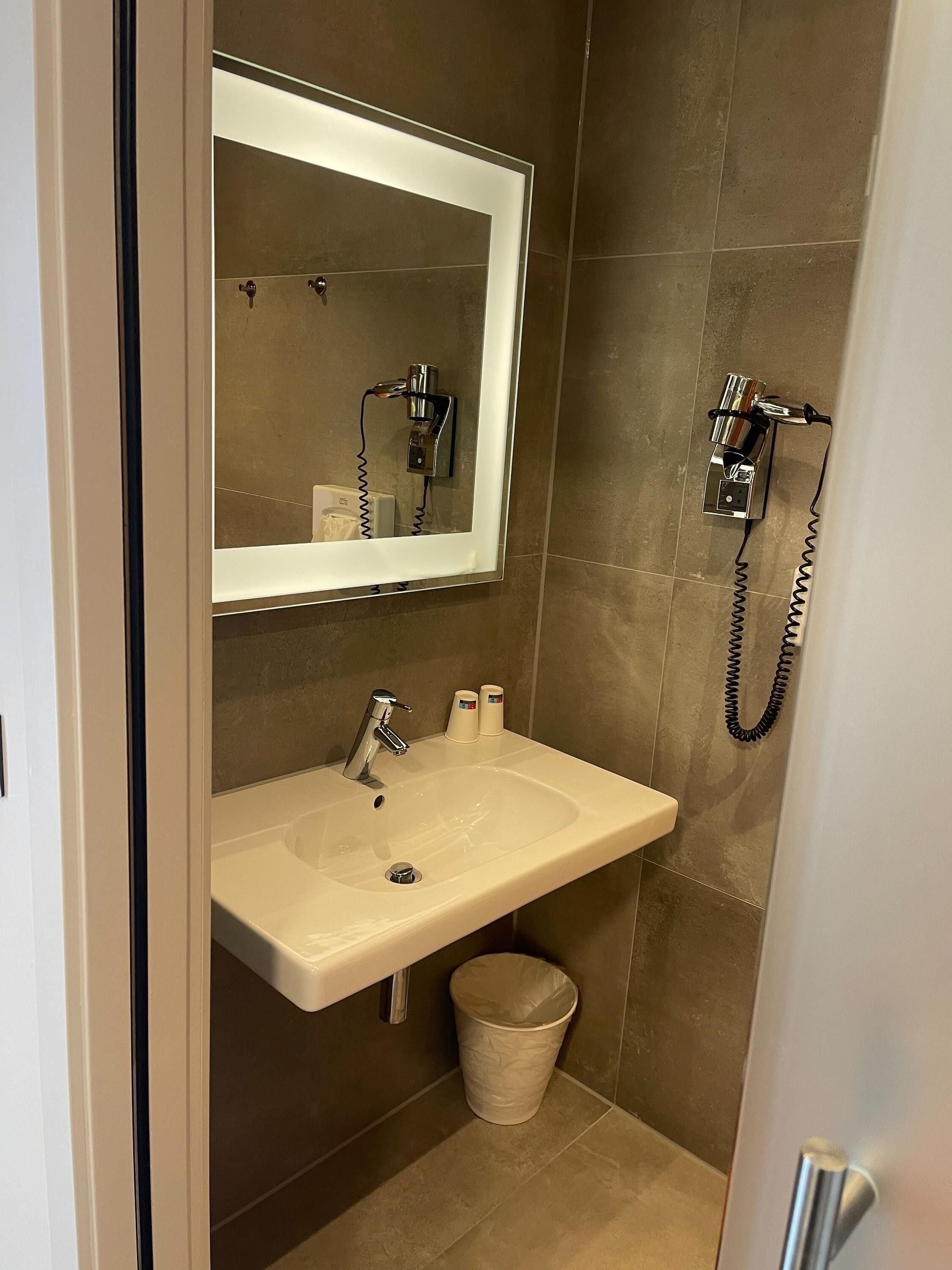 A bathroom with a white sink, a mirror, a hairdryer, and a trash bin.