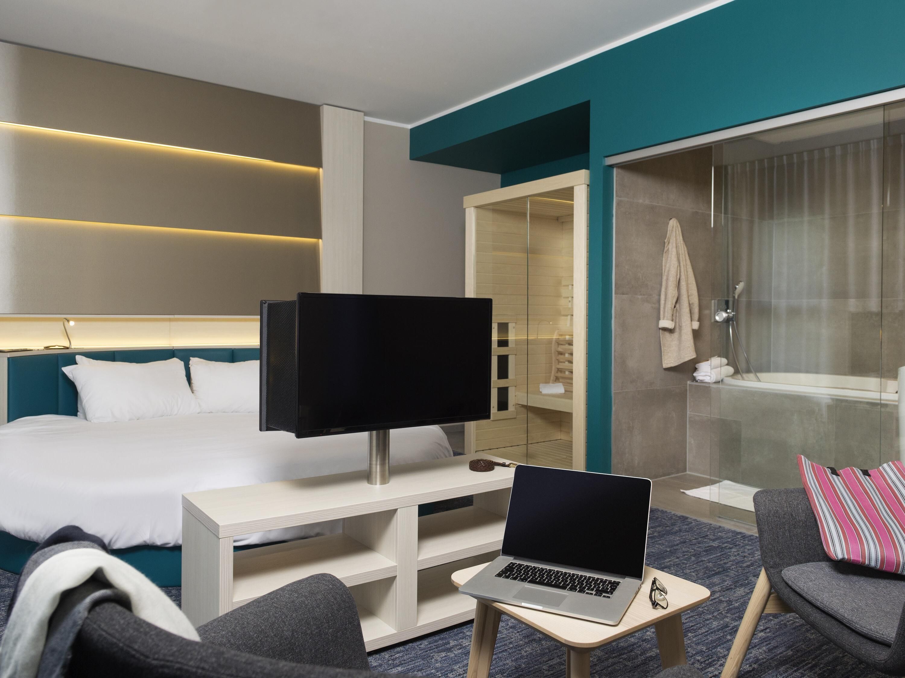 A hotel room with a bed, a desk with a laptop, a television, a shower area, and a seating area with a pillow.