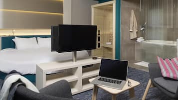A hotel room with a bed, a desk with a laptop, a television, a shower area, and a seating area with a pillow.
