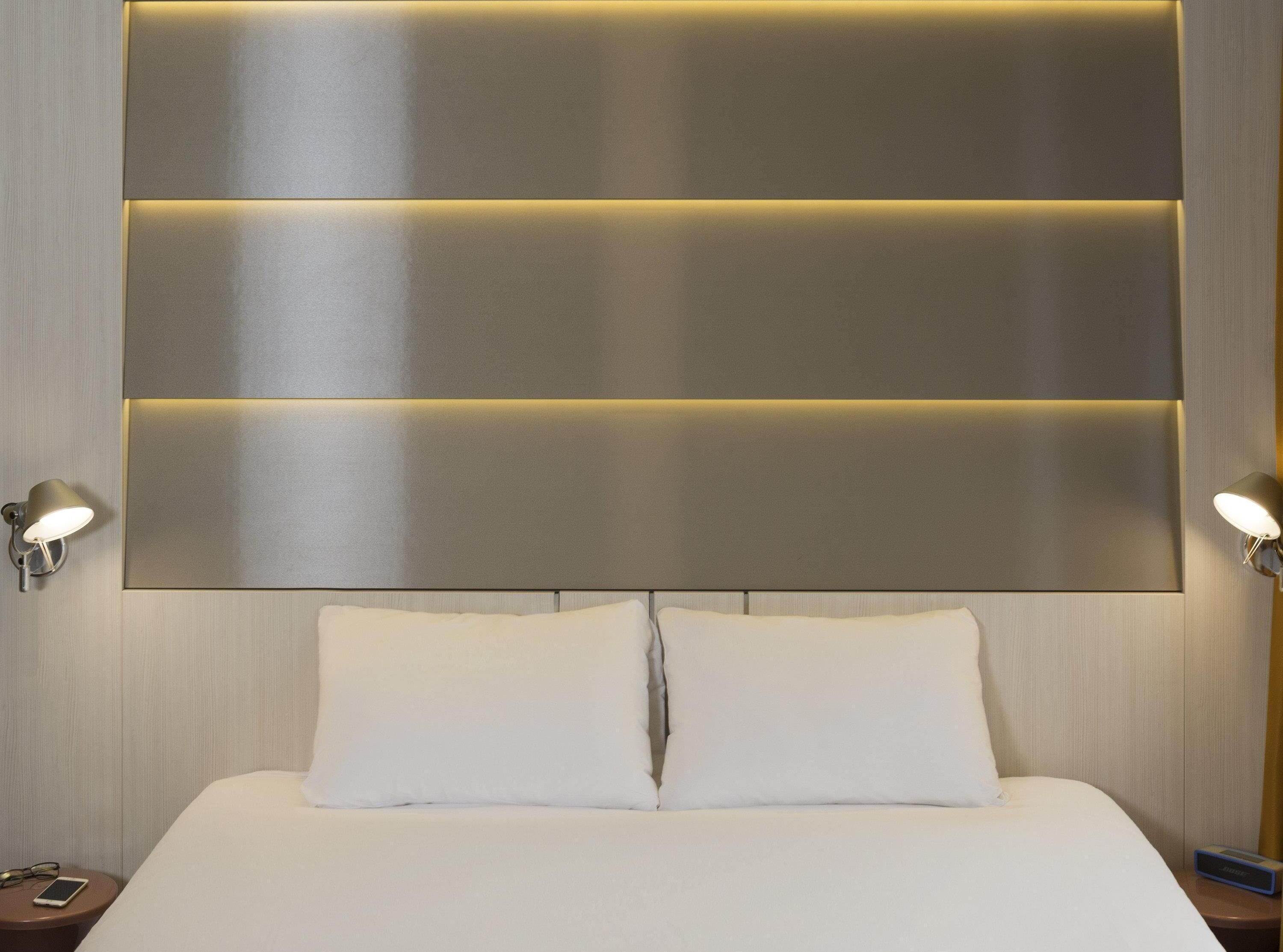Ein modernes Hotelzimmer mit einem großen Bett, zwei Nachttischlampen und einem an der Wand befestigten Kopfteil mit integrierter Beleuchtung.