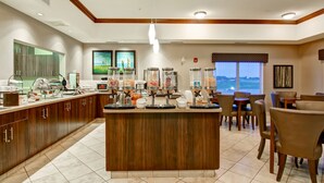 A modern hotel kitchen with a central island, breakfast bar, and dining area with chairs and tables.