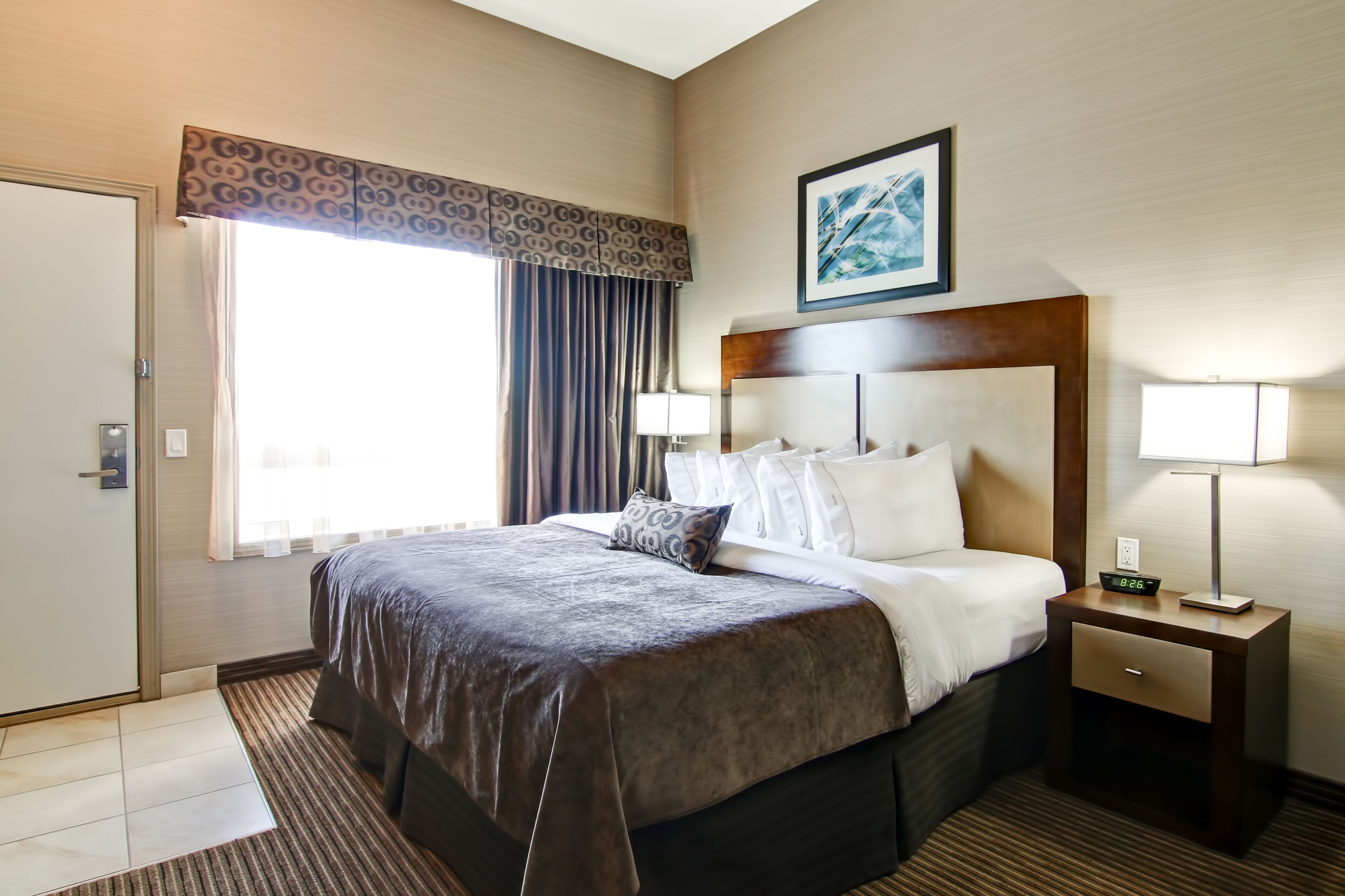 A hotel room with a large bed, a nightstand with a lamp, a window with curtains, and a painting on the wall.
