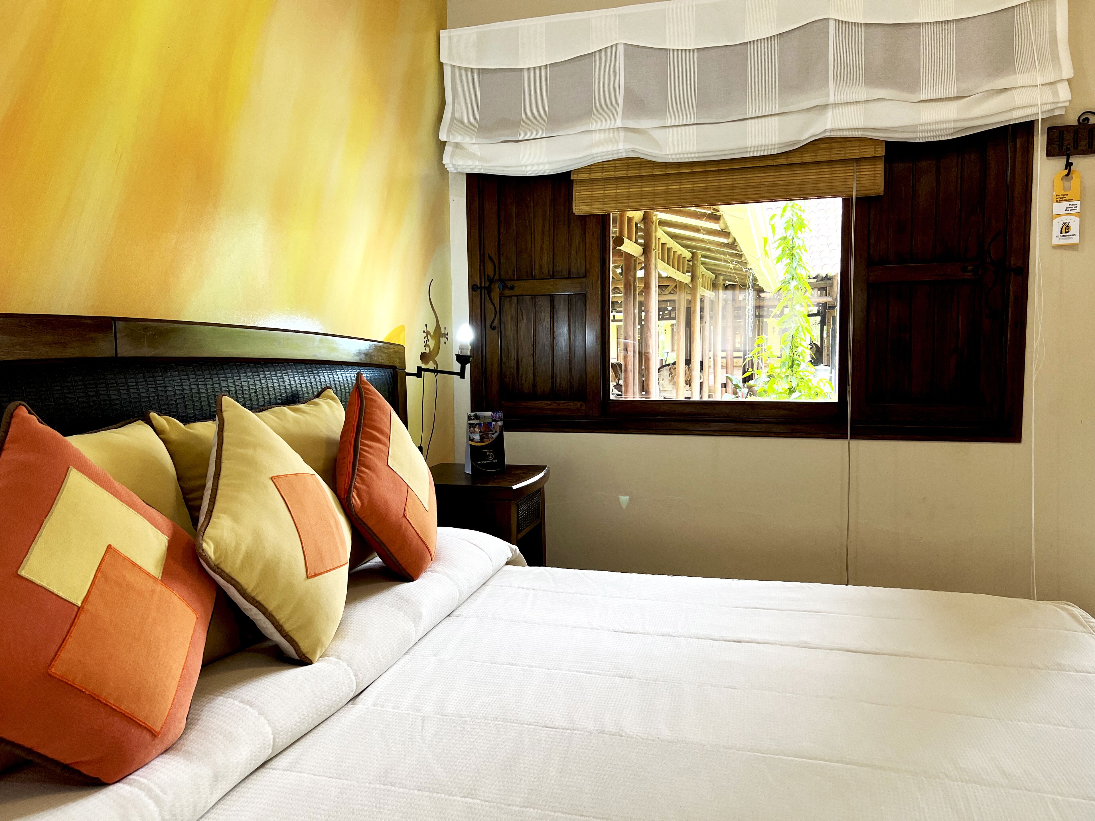 A hotel room with a bed, pillows, a nightstand, a lamp, a window with a view of a wooden structure, and a wall with a textured pattern.