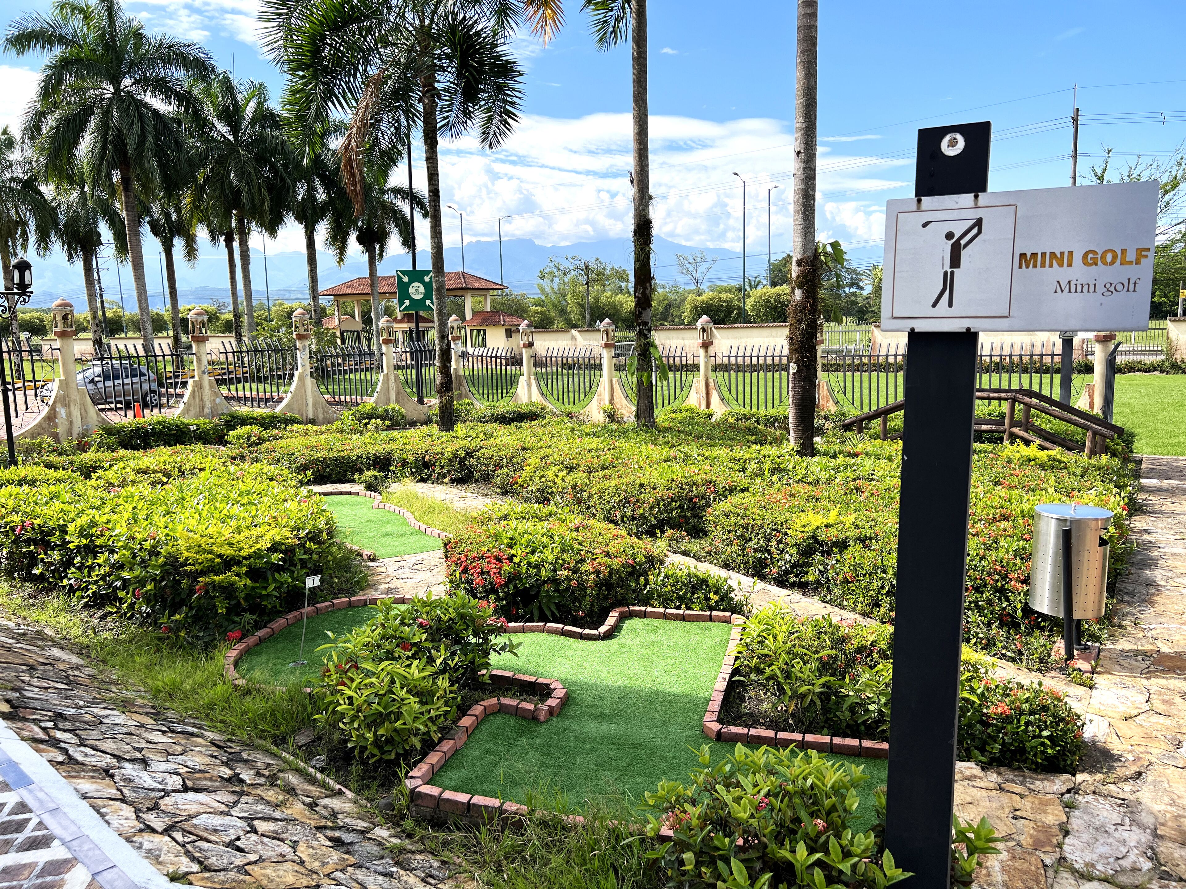 A mini golf course with a sign indicating its name, surrounded by well-maintained gardens and palm trees.