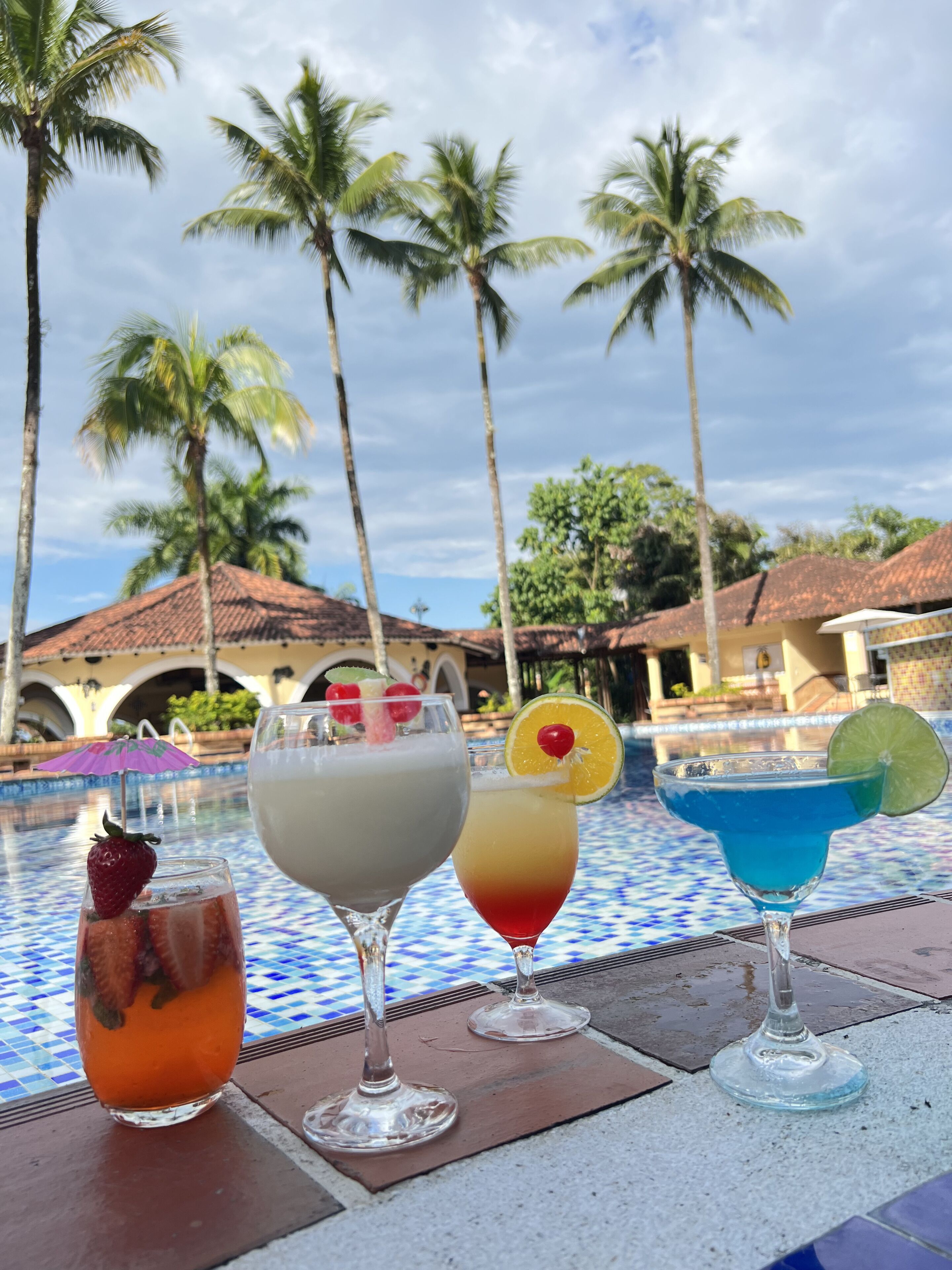 A poolside table with four drinks, palm trees, and a building with a tiled roof in the background.