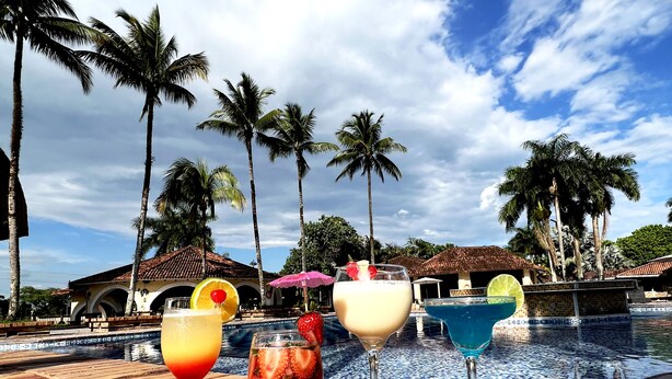 Zona junto a la piscina con tres bebidas, palmeras y una piscina con baldosas.