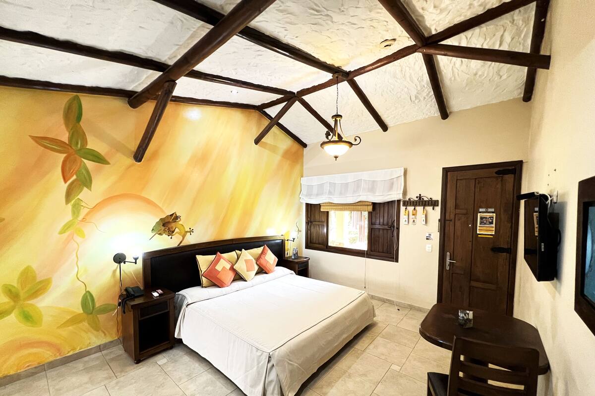 A bedroom with a bed, nightstands, a desk, and a chair. There is a window with shutters and a wall-mounted TV.
