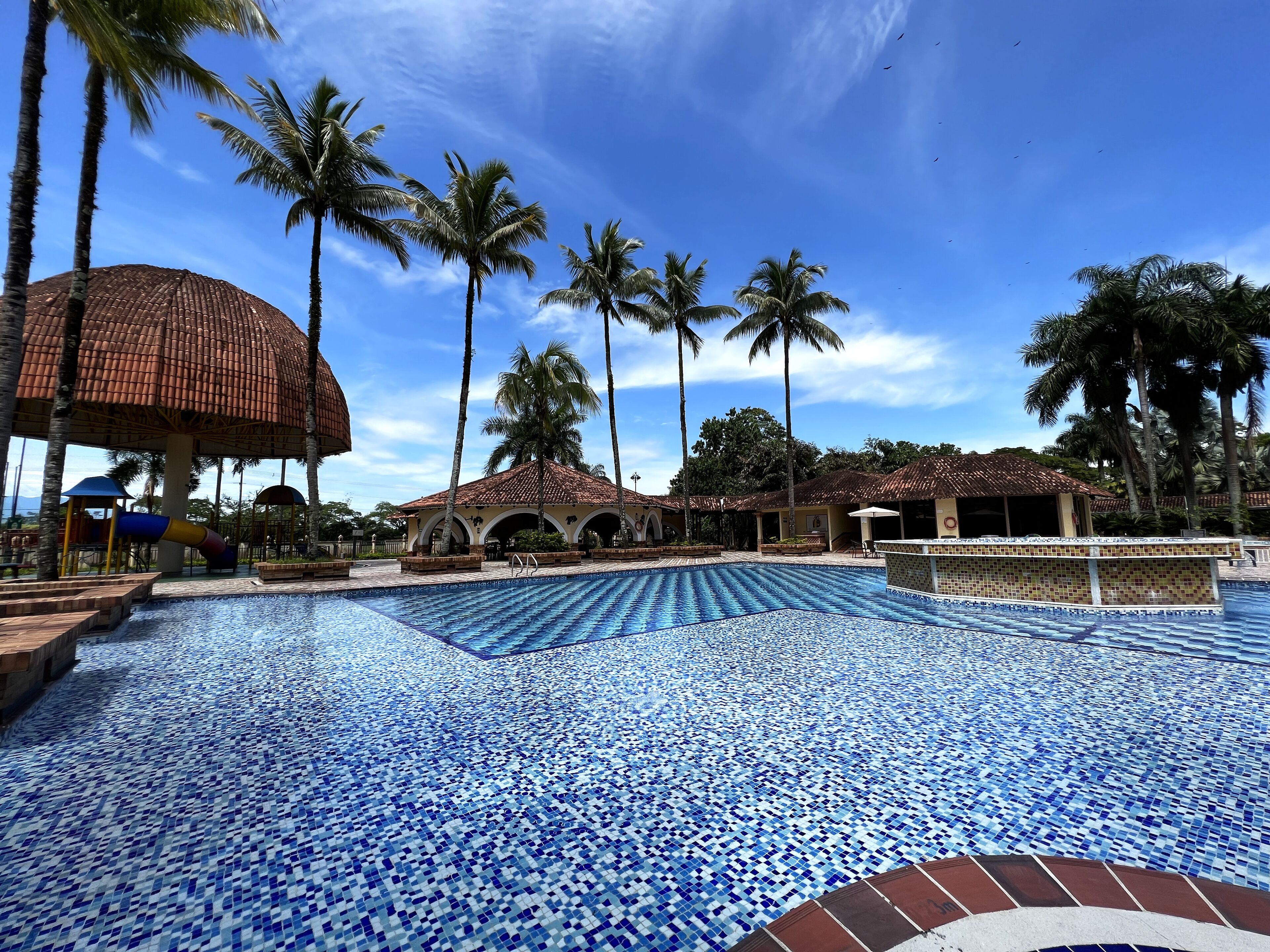 A swimming pool with blue mosaic tiles, surrounded by palm trees and a building with a tiled roof.