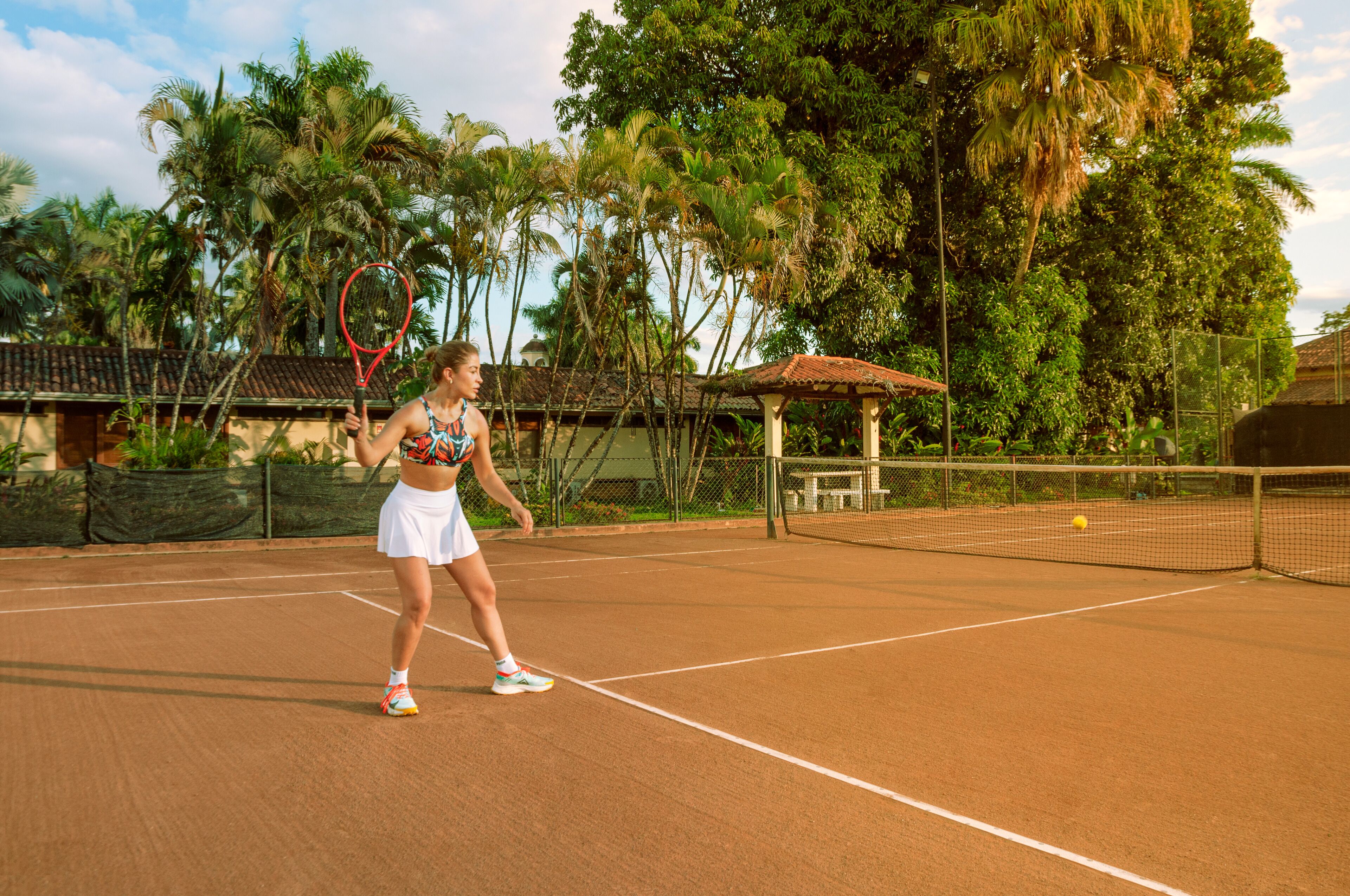 A tennis court with a net, surrounded by palm trees and a small pavilion.