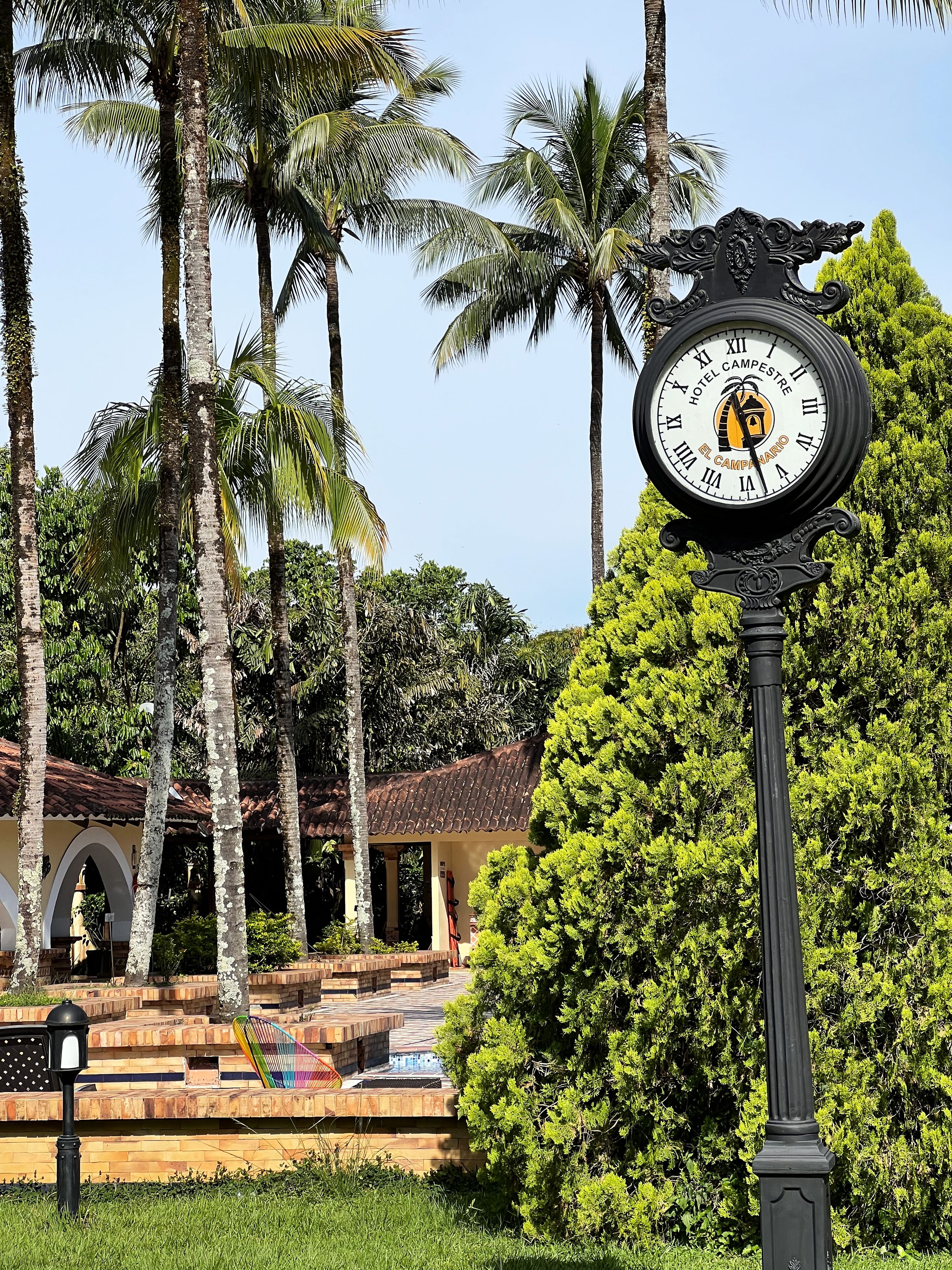 A clock with a hotel logo, surrounded by palm trees and a building with a tiled roof.