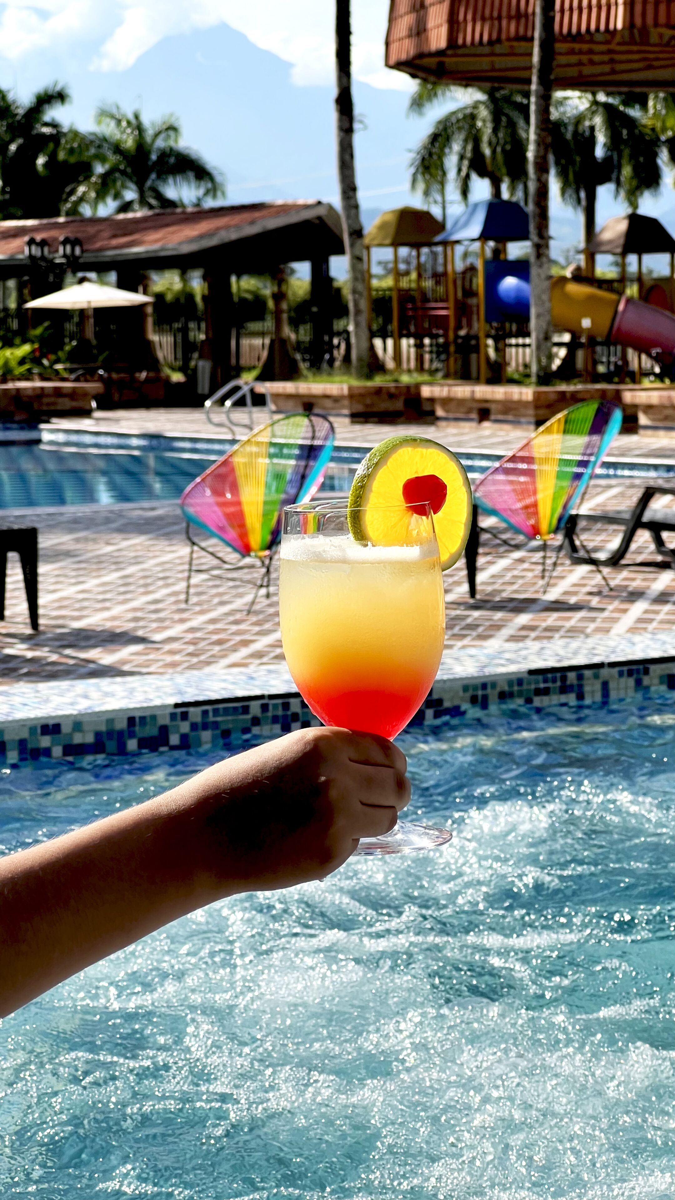 A hand holding a cocktail with a lime wedge and a cherry, near a pool with lounge chairs and palm trees in the background.