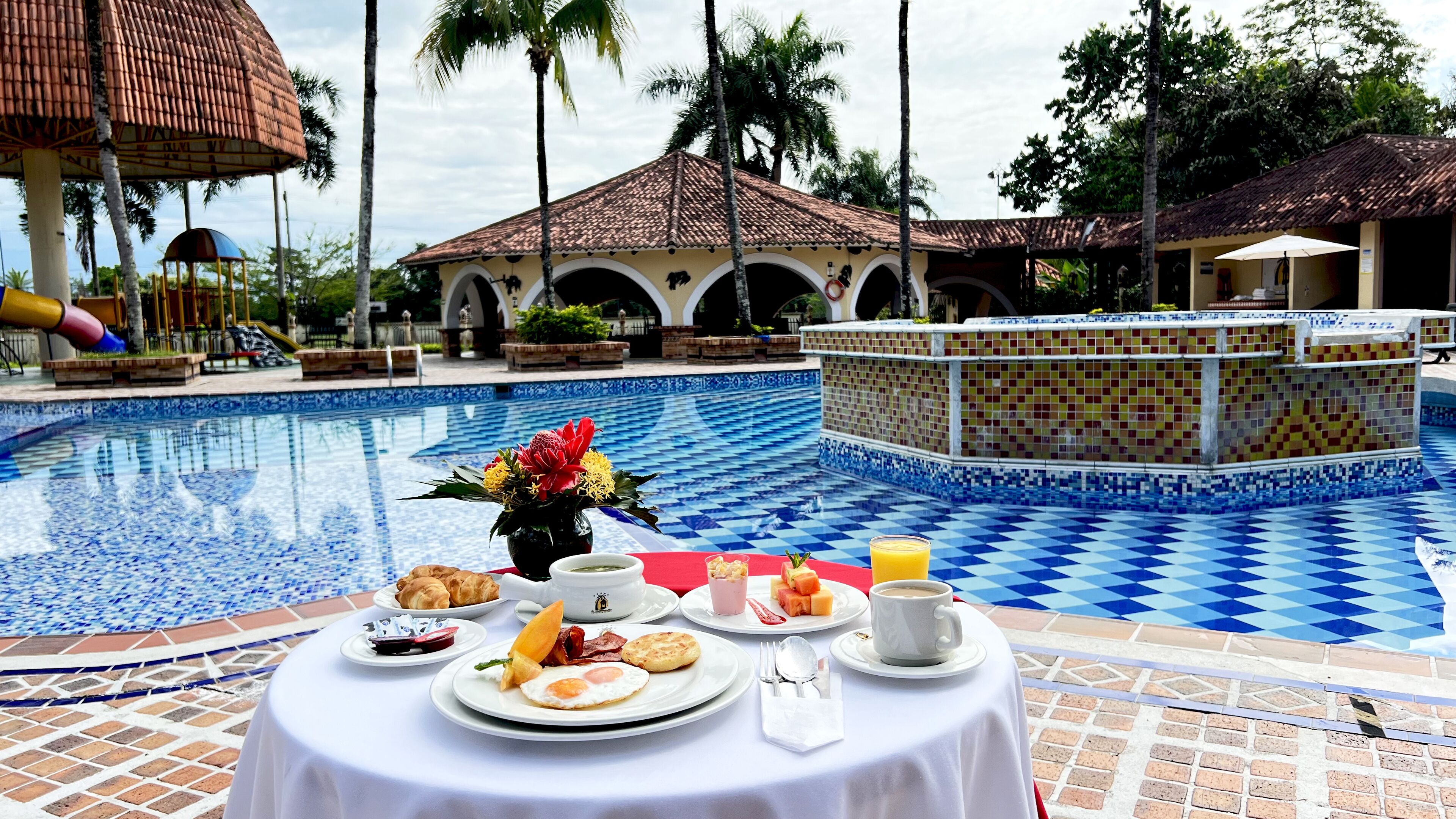 A poolside breakfast setup with a table, plates of food, and a drink.