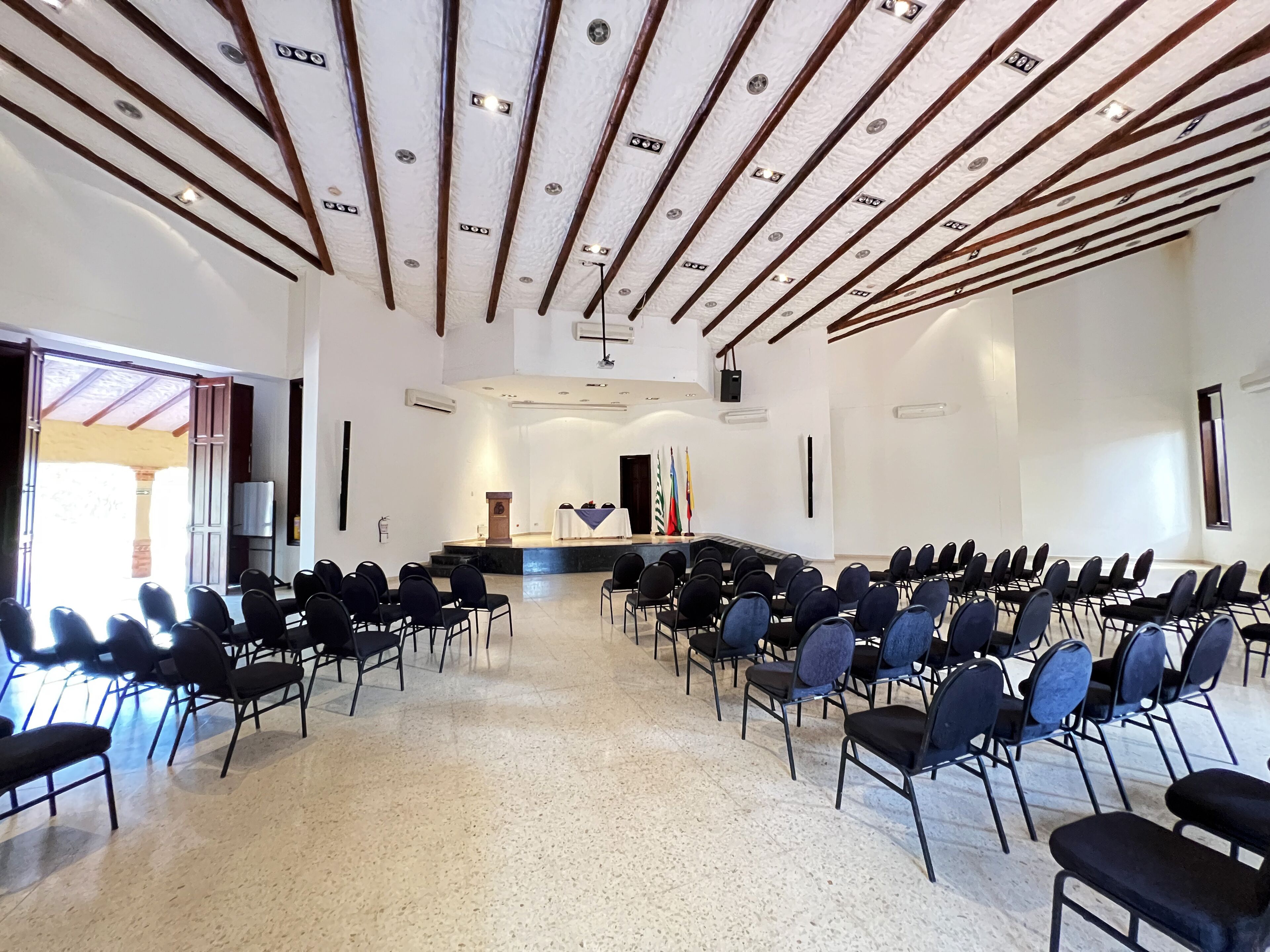 Un salón de conferencias con filas de sillas orientadas hacia un escenario con un atril y banderas.