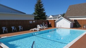 A rectangular swimming pool with a wooden deck surrounding it. There are white lounge chairs and a wooden fence in the background. - Bridge View Motel & Condo's (Mackinaw City)