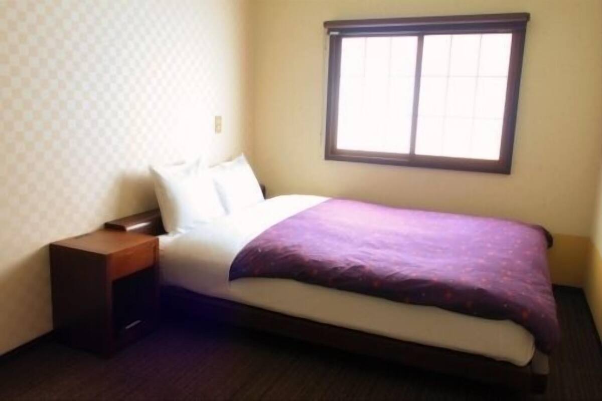 Una cama individual con estructura de madera, una mesita de noche, una ventana con persianas y un papel tapiz estampado.