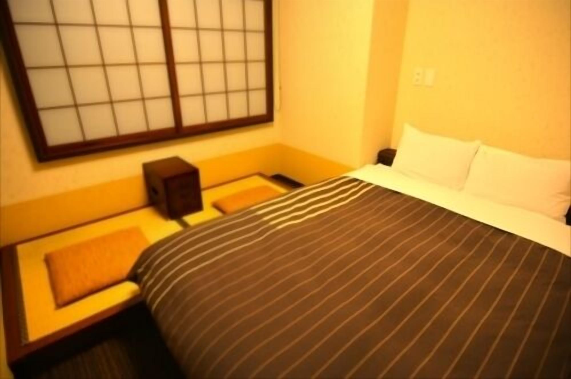 Ein Hotelzimmer mit Bett, Nachttisch, kleinem Tisch, einem Fenster mit Jalousien und einem wandmontierten Lichtschalter.