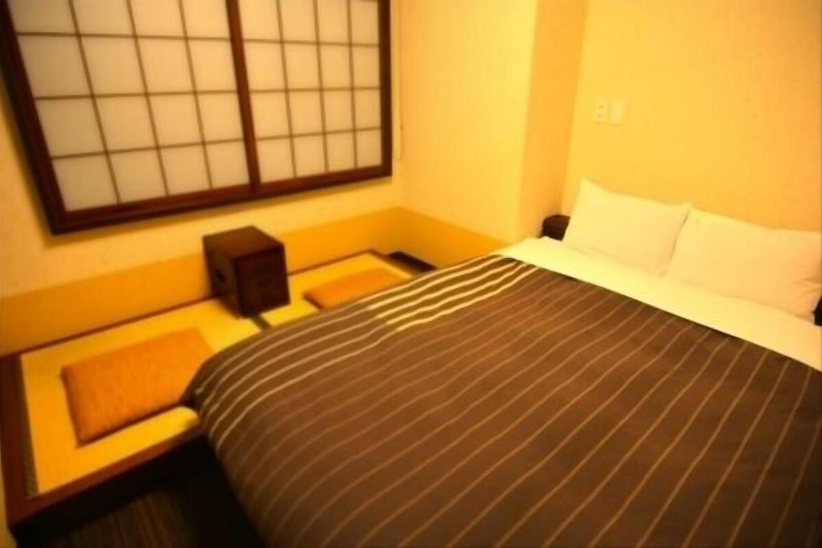 A hotel room with a bed, a nightstand, a small table, a window with blinds, and a wall-mounted light switch.