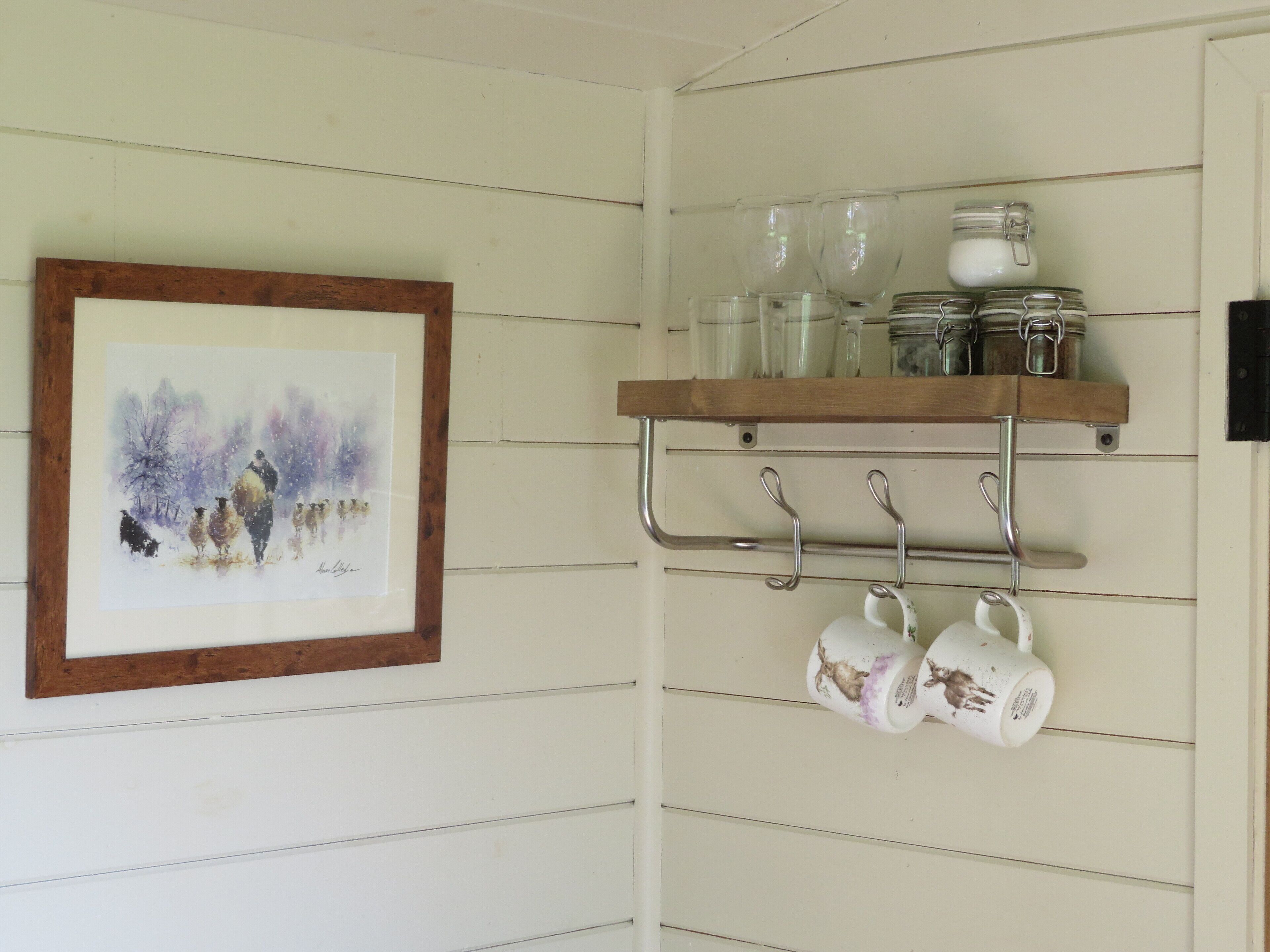 Une cuisine avec une étagère en bois où sont posés des bocaux et des tasses, et un tableau encadré accroché au mur.