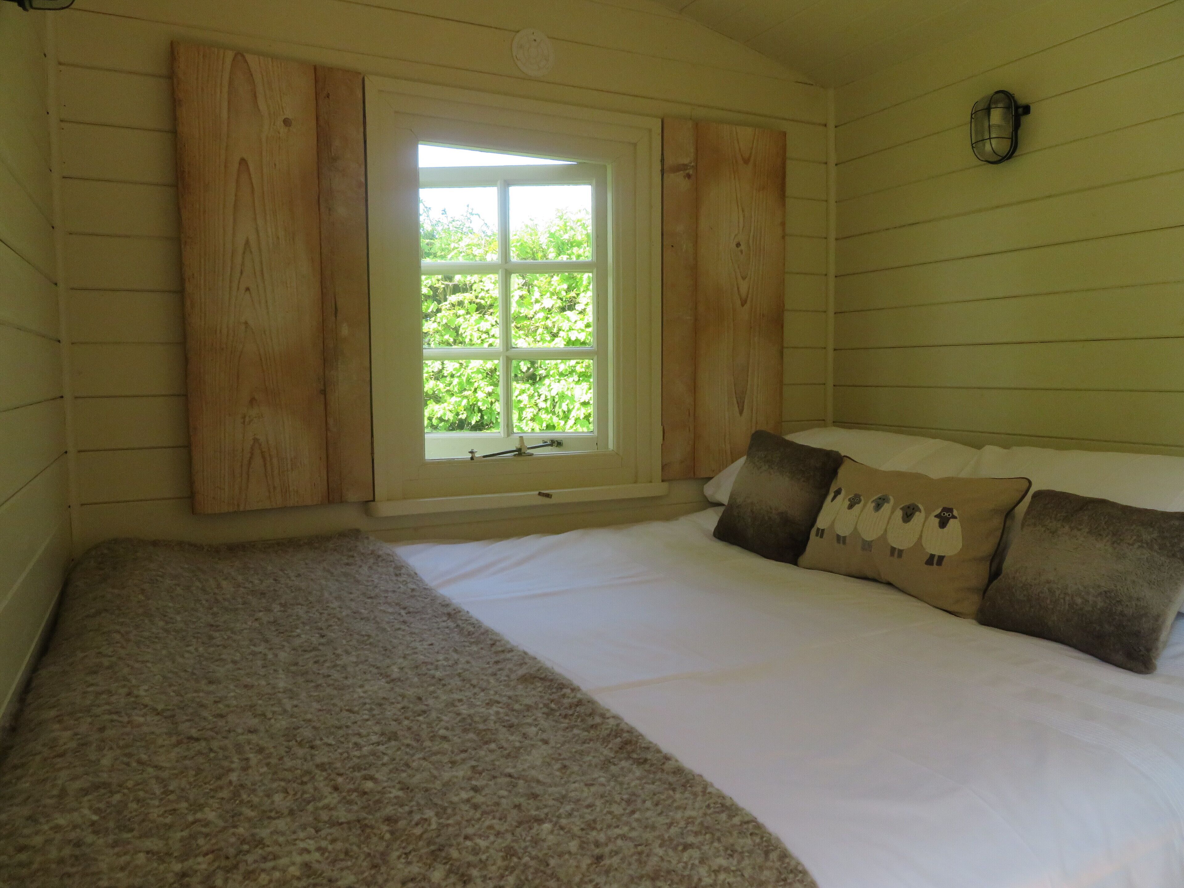 A bedroom with a large window, wooden panel walls, a bed with white bedding and two pillows, and a small wall-mounted light fixture.