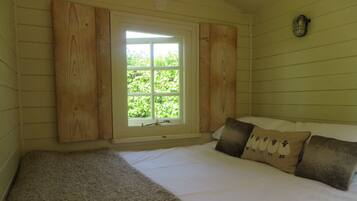 A bedroom with a large window, wooden panel walls, a bed with white bedding and two pillows, and a small wall-mounted light fixture.