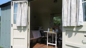 Une petite cabane d’une seule pièce, avec un plancher en bois, un lit, une table avec des chaises et une fenêtre ornée de rideaux à motifs.