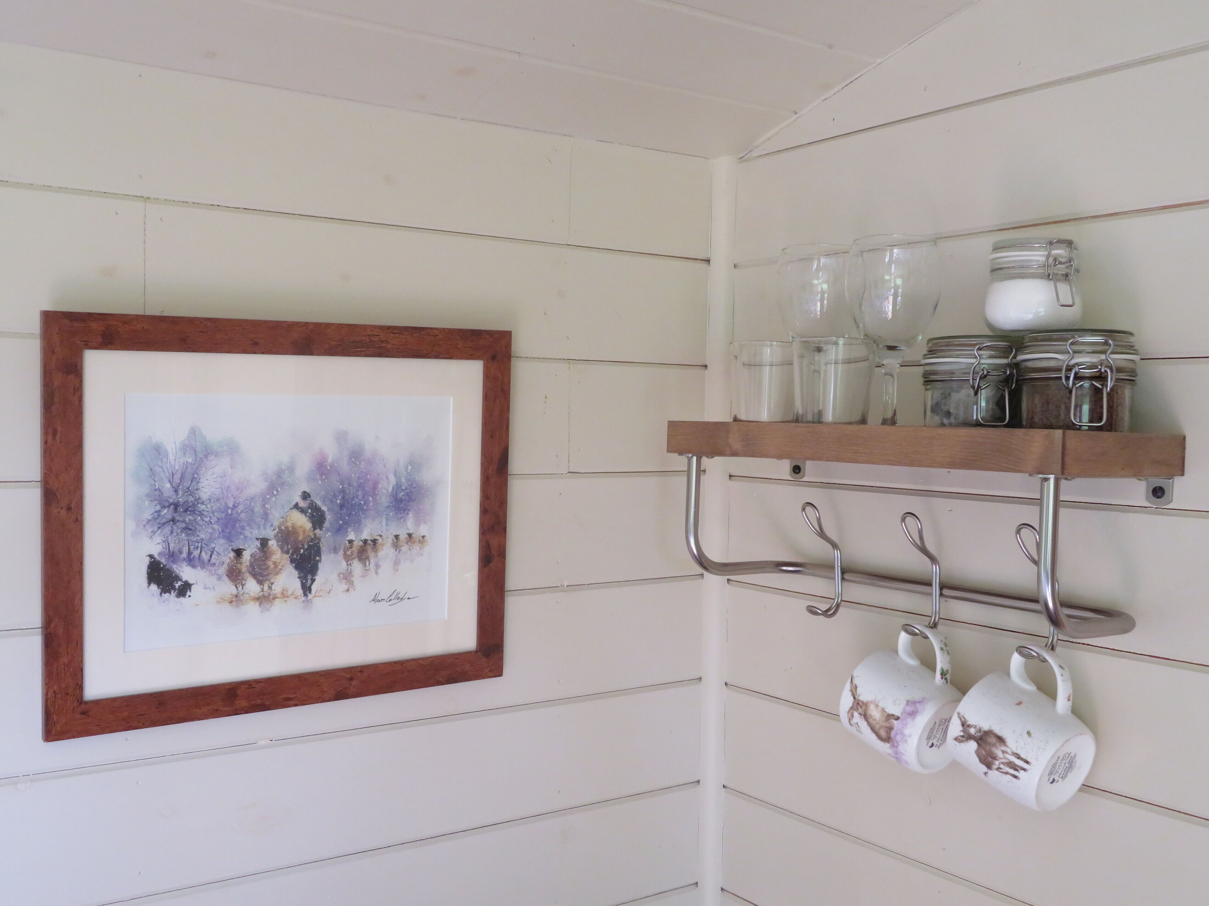 A kitchen with a wooden shelf holding jars and mugs, and a framed painting on the wall.