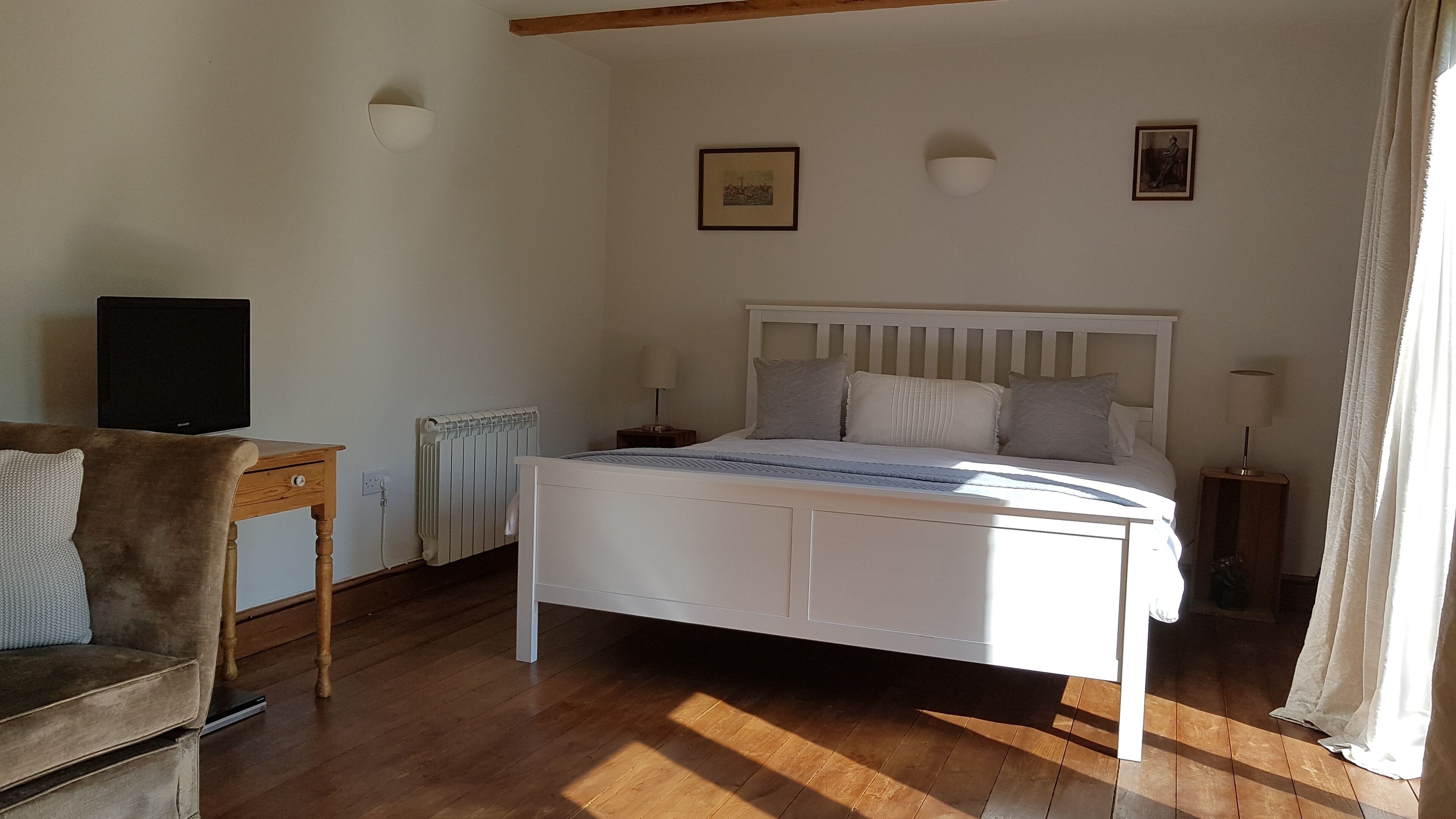 Quarto com cama branca, piso de madeira, televisão em uma mesa e uma cadeira.