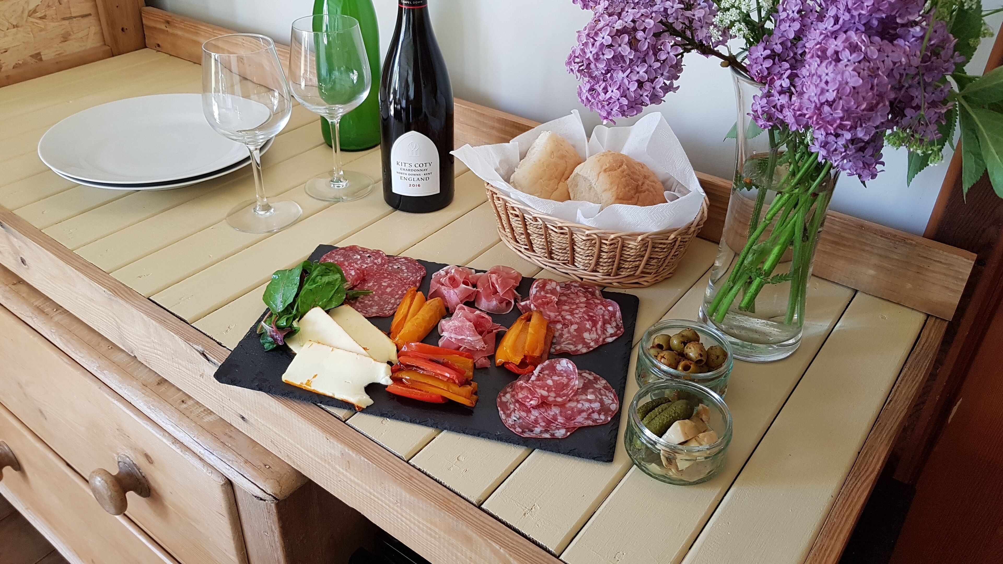Ett träbord med en svart skifferplatta där olika sorters ostar, lufttorkat kött och oliver är uppradade. En vinflaska, ett vinglas och en korg med brödbullar syns också.