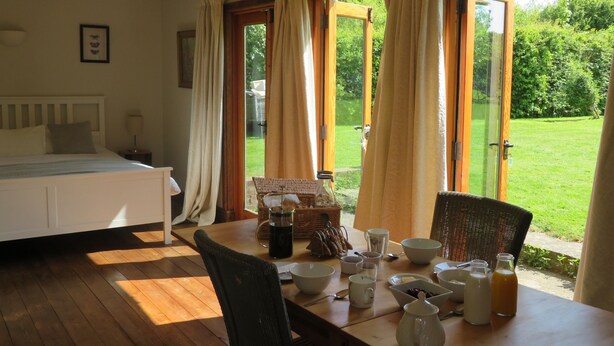 Une pièce comprenant un lit, une table à manger avec divers objets, et donnant sur un jardin grâce à des portes ouvertes.