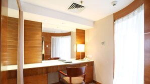 A hotel room with a wooden desk, a chair, a large mirror, a lamp, and a window with curtains.