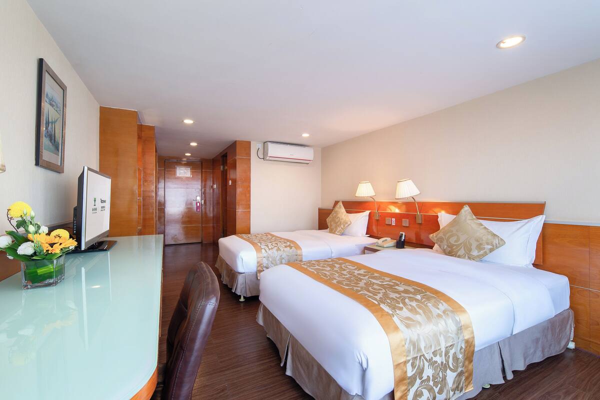 Ein Hotelzimmer mit zwei Betten, einem Schreibtisch mit Computer und einer Blumenvase sowie einer Holzwandverkleidung.