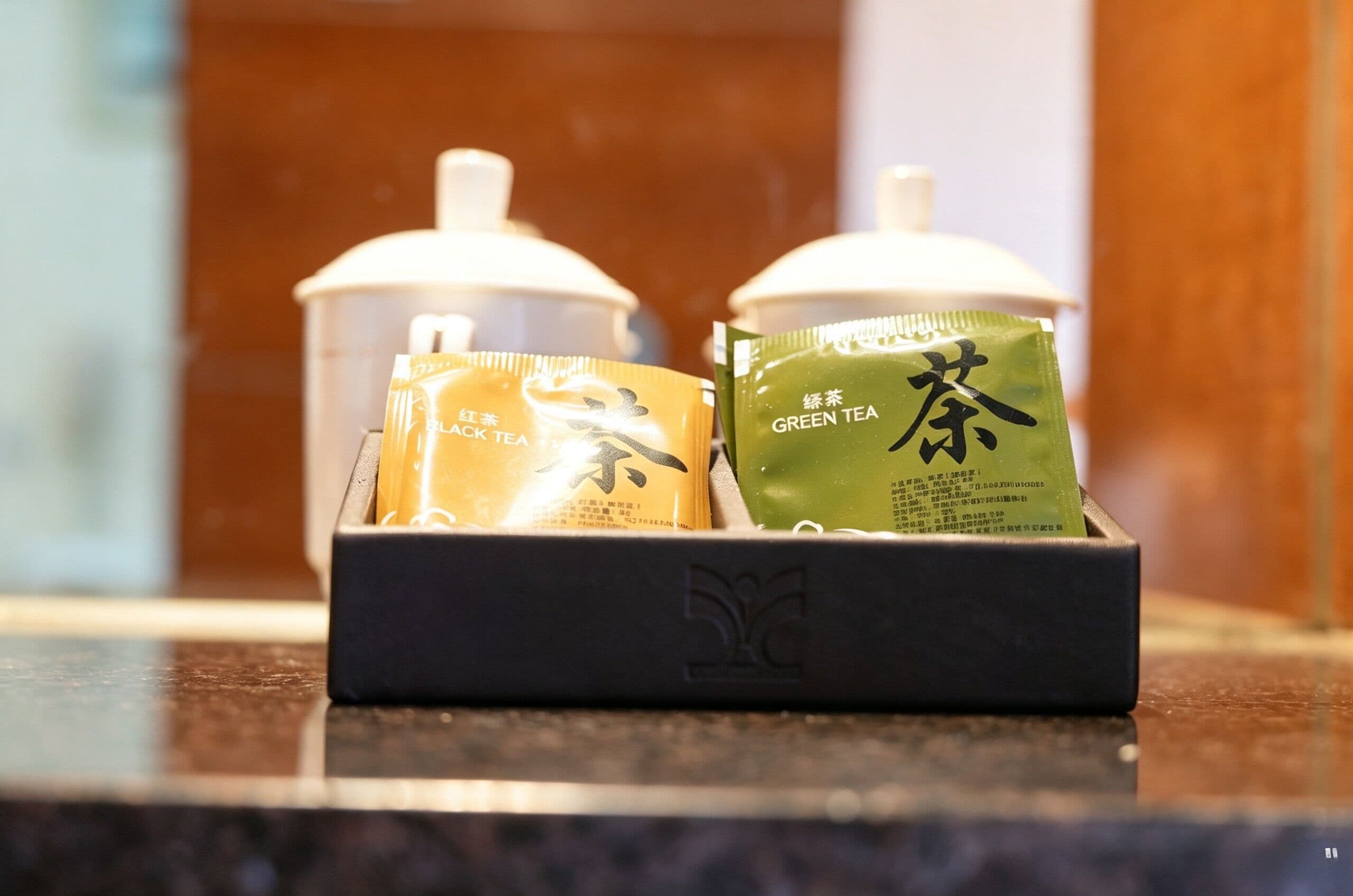 Two containers with tea packets, one labeled 'BLACK TEA' and the other 'GREEN TEA', placed on a counter.